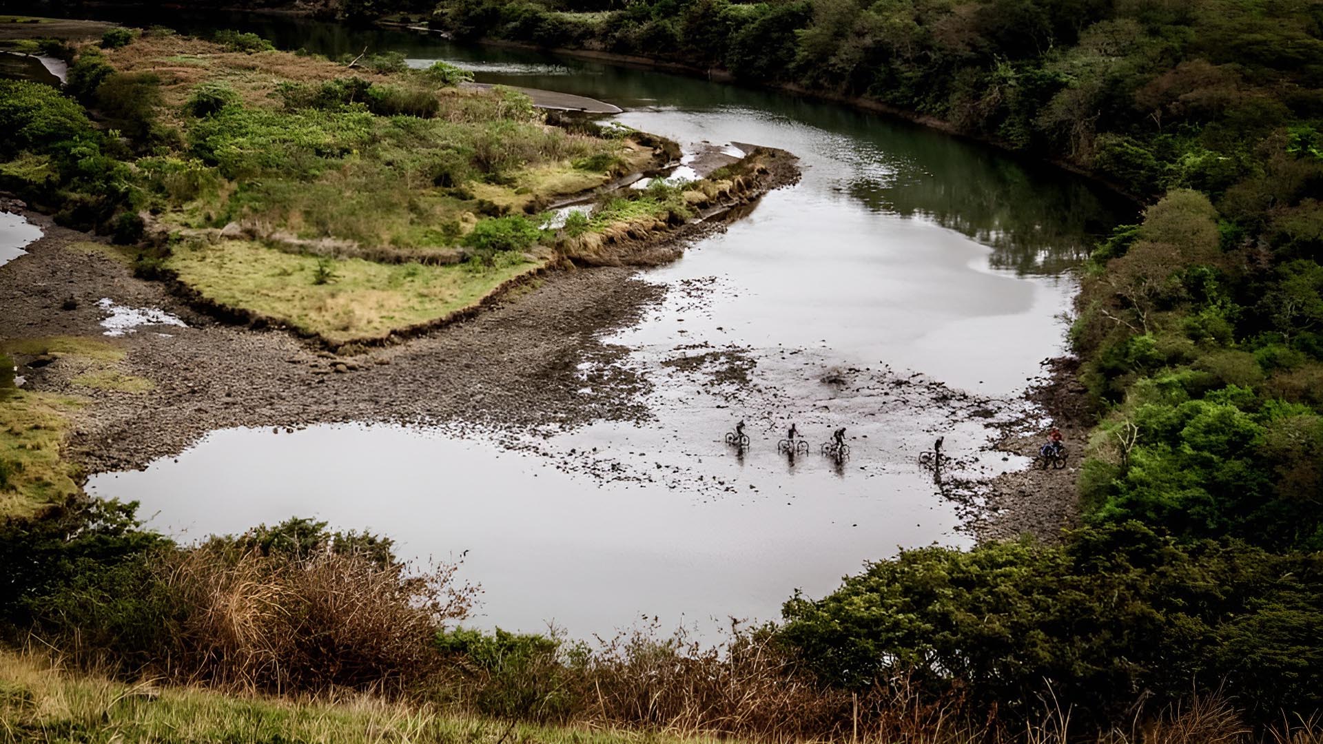 Adventurers on mountain bikes riding through mud to cross a shallow river.