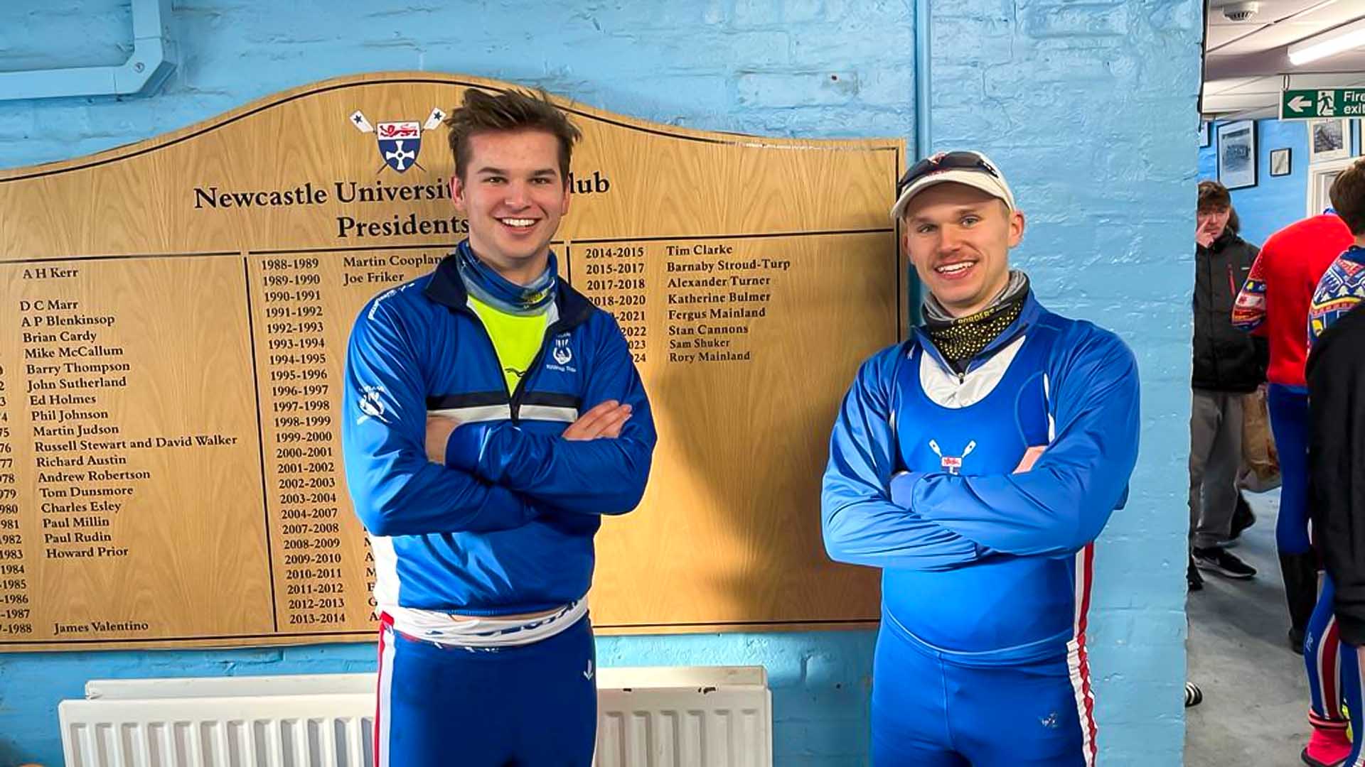 Two brothers posing next to the President's Board at Newcastle University Boat Club.