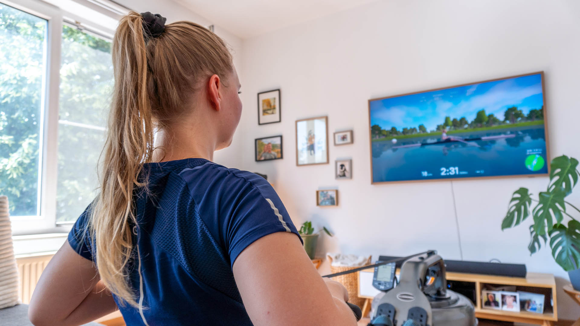 Young woman rowing on her rowing machine in the living room while training with the virtual rowing app EXR.