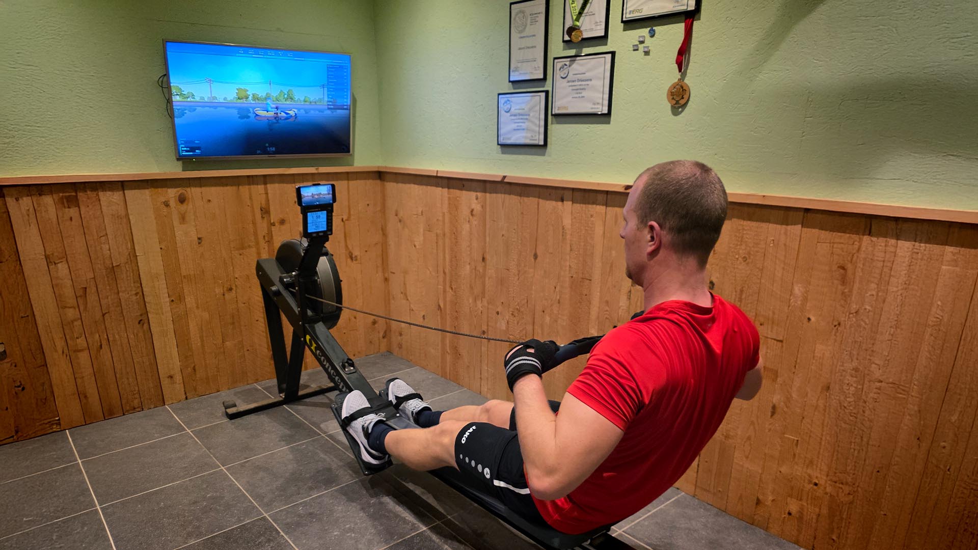 Man rowing on his Concept2 rowing machine in his home gym with EXR on a flatscreen TV mounted to the wall.