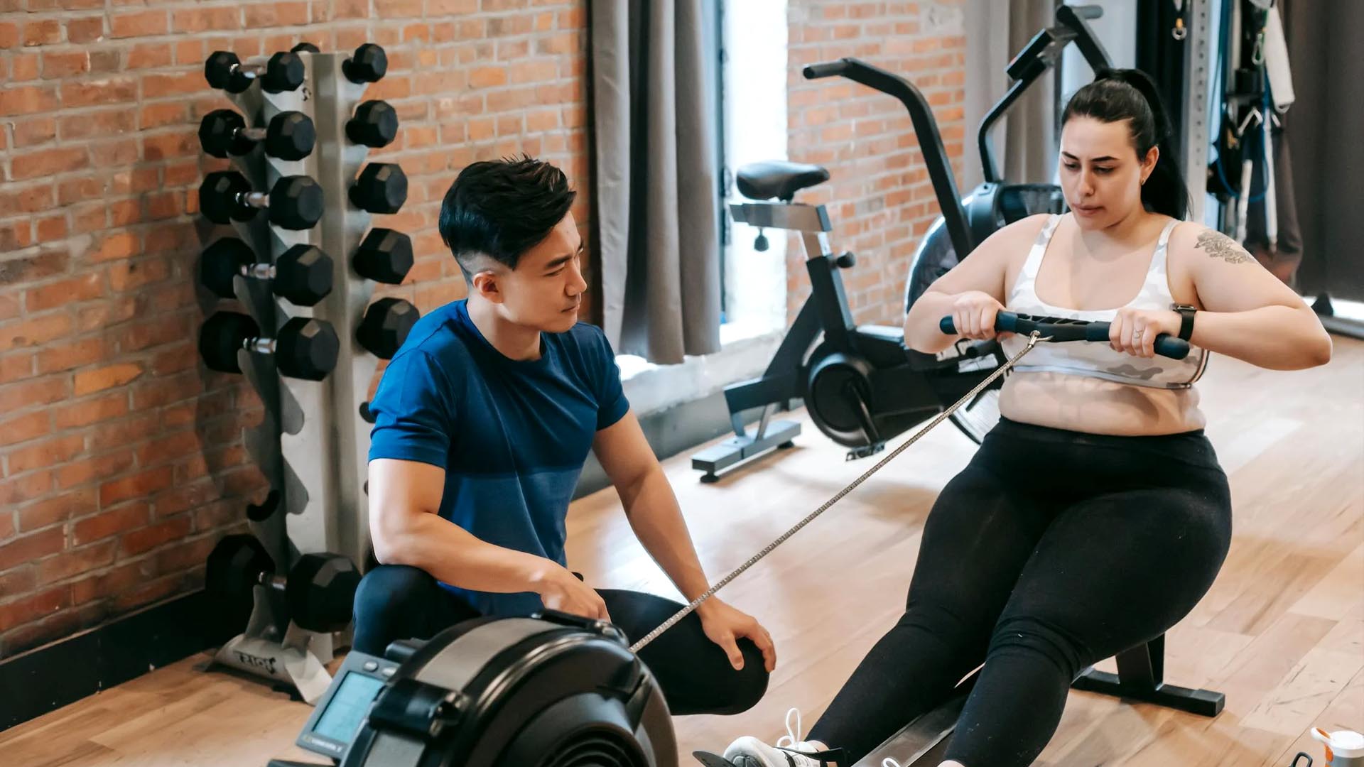 A Woman learning how to use the indoor rowing machine with the help of a personal trainer.