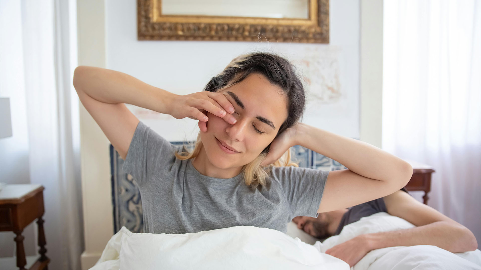 Woman rubbing her eyes in her bed while her partner is sleeping next to her.